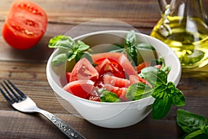 Fresh red tomatoes are sliced in a white ceramic plate with basil herb and olive oil on a wooden table