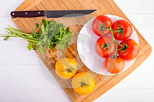 Fresh red and yellow tomatoes from above on a wooden board with a knife in a white plate at a white table