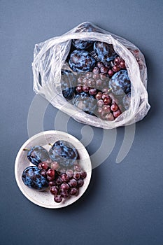 Fresh ripe plum fruits and grape berries in plastic bag package and in wooden bowl on minimal blue grey background