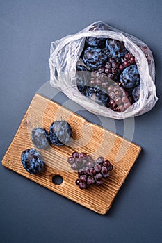 Fresh ripe plum fruits and grape berries in plastic bag package and on wooden cutting board on minimal blue grey background