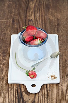 Fresh ripe red strawberries in blue enamel mug on white ceramic board over rustic wooden background