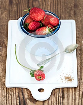 Fresh ripe red strawberries in blue enamel mug on white ceramic board over rustic wooden background