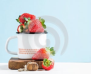 Fresh ripe red strawberries in country style enamel mug on rustic wooden board, pastel light blue background