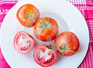 Fresh ripe ripe red tomatoes and cut tomato with water drops and green peduncle on white plate - top view, organic vegetables