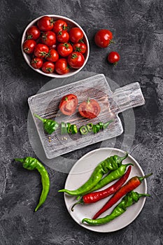 Fresh ripe tomatoes in bowl near to old wooden cutting board with hot chili peppers in plate, dark stone concrete background