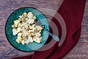 Fresh roasted cauliflower in a blue-green dinner bowl on a rustic wood table, black fork, red kitchen towel