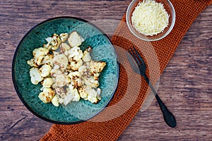 Fresh roasted cauliflower in a blue-green dinner bowl on a rustic wood table, small bowl parmesan cheese, black fork