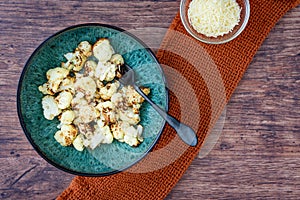 Fresh roasted cauliflower in a blue-green dinner bowl on a rustic wood table, small bowl parmesan cheese, black fork, orange kitch