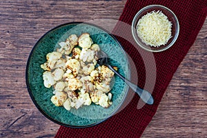 Fresh roasted cauliflower in a blue-green dinner bowl on a rustic wood table, small bowl parmesan cheese, black fork, red kitchen