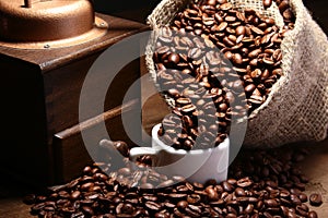 Fresh roasted coffee beans in burlap sack, coffee cup and grinder on dark background.