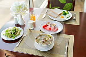 Fresh romantic breakfast table next to window with bread, fruit, juice, milk, noodle soup