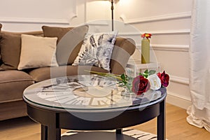 Fresh roses on coffee table with clock inside. Interior of living room with cosy sofa.