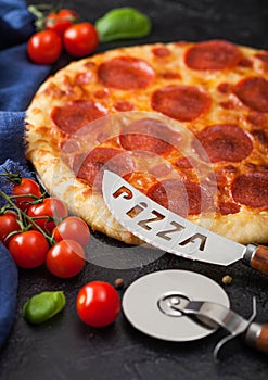 Fresh round baked Pepperoni italian pizza with wheel cutter and knife with tomatoes and basil on black background. Close up