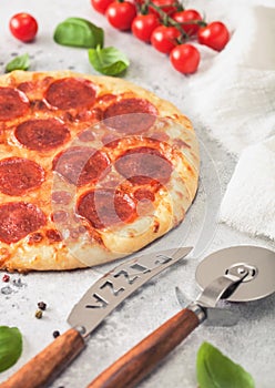 Fresh round baked Pepperoni italian pizza with wheel cutter and knife with tomatoes and basil on light background with linen towel