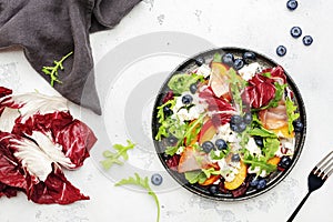 Fresh salad with arugula, radicchio; peaches, smoked pork ham, feta cheese and blueberries. White table background, top view