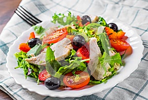 Fresh salad with chicken breast, arugula, black olives,red pepper, lettuce, fresh sald leaves and tomato on a white plate on woode