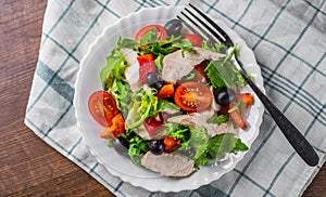 Fresh salad with chicken breast, arugula, black olives,red pepper, lettuce, fresh sald leaves and tomato on a white plate on woode