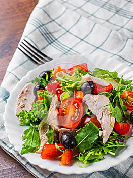 Fresh salad with chicken breast, arugula, black olives,red pepper, lettuce, fresh sald leaves and tomato on a white plate on woode