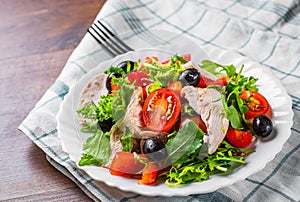 Fresh salad with chicken breast, arugula, black olives,red pepper, lettuce, fresh sald leaves and tomato on a white plate on woode