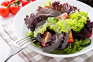 Fresh salad with green and purple lettuce, tomatoes and cucumbers on white wooden background close up.