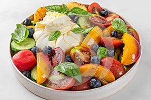 Fresh salad with grilled peaches, burrata cheese, blueberries, vegetables and basil in a plate on white background