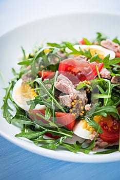 Fresh salad with tuna, tomatoes, eggs, arugula and mustard on blue wooden background close up