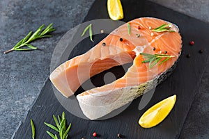 Fresh salmon steak with ingredients for cooking on a board on dark background. Raw trout steak with salt, pepper, lemon and