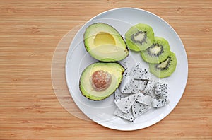 Fresh sliced fruit avocado, dragon fruit and kiwi on white plate against wooden board background with copy space