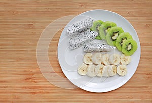 Fresh sliced fruit banana, dragon fruit and kiwi on white plate against wooden board background with copy space