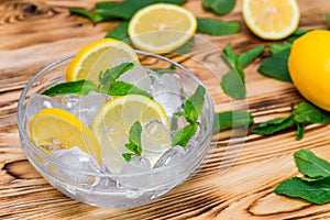 Fresh sliced lemon, bright green mint leaves and frozen ice cubes in a transparent bowl on a wooden table