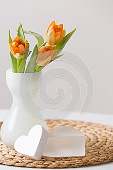 Fresh spring bunch of orange tulips in a nice white glass vase and two cute heart symbols on the straw board. Home decor