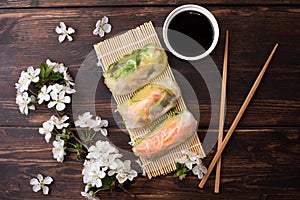 Fresh spring rolls with soy sauce, chopsticks and cherry blossom sprig on dark wooden background, asian food