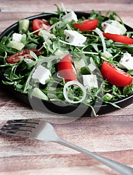 Fresh spring salad with arugula, feta cheese, red onion and tomatoes in a black bowl.
