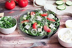 Fresh spring salad with arugula, feta cheese, red onion and tomatoes in a black bowl.