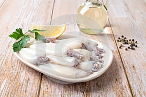 Fresh squid tubes on a plate over wooden table. Small calamary fillet, lemon and olive oil prepared for cooking. Raw squid for low