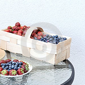 Fresh strawberries and blueberries lie in a plate and basket on a glass table against the background of a plastered white wall. Fo