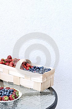 Fresh strawberries and blueberries lie in a plate and basket on a glass table against the background of a plastered white wall.