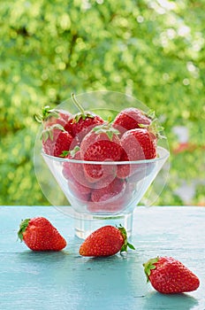 Fresh strawberries in a glass dessert bowl on the blue kitchen table on the blurred nature background. Vegan summer breakfast