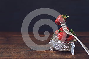 Fresh strawberries in metal plate and one berry on fork still life on dark wooden background. Organic garden strawberry on rustic