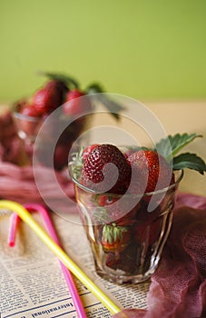 Fresh strawberry in two glasses with cocktail tubes on yellow table with newspaper napkin and green background, summer drink