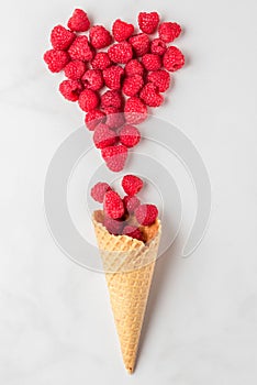 Fresh summer raspberries in waffle ice cream cone with heart made of berries on white marble background