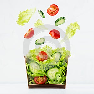 Fresh summer salad of raw vegetable with flying ingredients - tomatoes, cucumber, green salad above craft box for take away food.