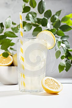 Fresh summer sour lemonade in glass with ice, straw, lemon slice in elegant modern kitchen with white table, marble tile wall.