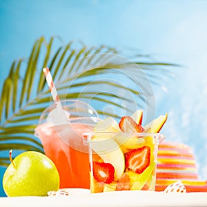 Fresh summer takeaway food on tropical beach - fruit salad, cold drink, green apple, sun hat on white sand with palm leaves.