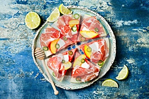 Fresh summer watermelon pizza with feta cheese, peach, prosciutto, jalapeno and honey drizzle on blue background.