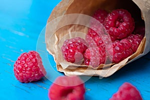 Fresh tasty raw red raspberries in the paper cone on blue painted background with wood texture.