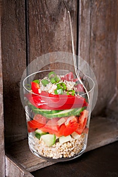 Fresh tasty vegetarian or vegan layer salad with tomatoes, cucumber, paprika, green onion and grain in a glass on a wooden