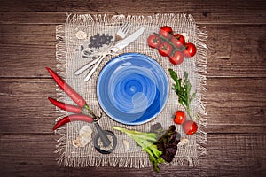 Fresh tomatoes, chili pepper and other spices and herbs around modern dark blue plate in the center of wooden table and cloth