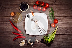 Fresh tomatoes, chili pepper and other spices and herbs around modern white square plate in the center of wooden table.