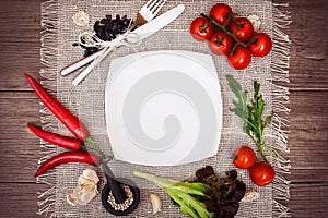 Fresh tomatoes, chili pepper and other spices and herbs around modern white square plate in the center of wooden table and cloth